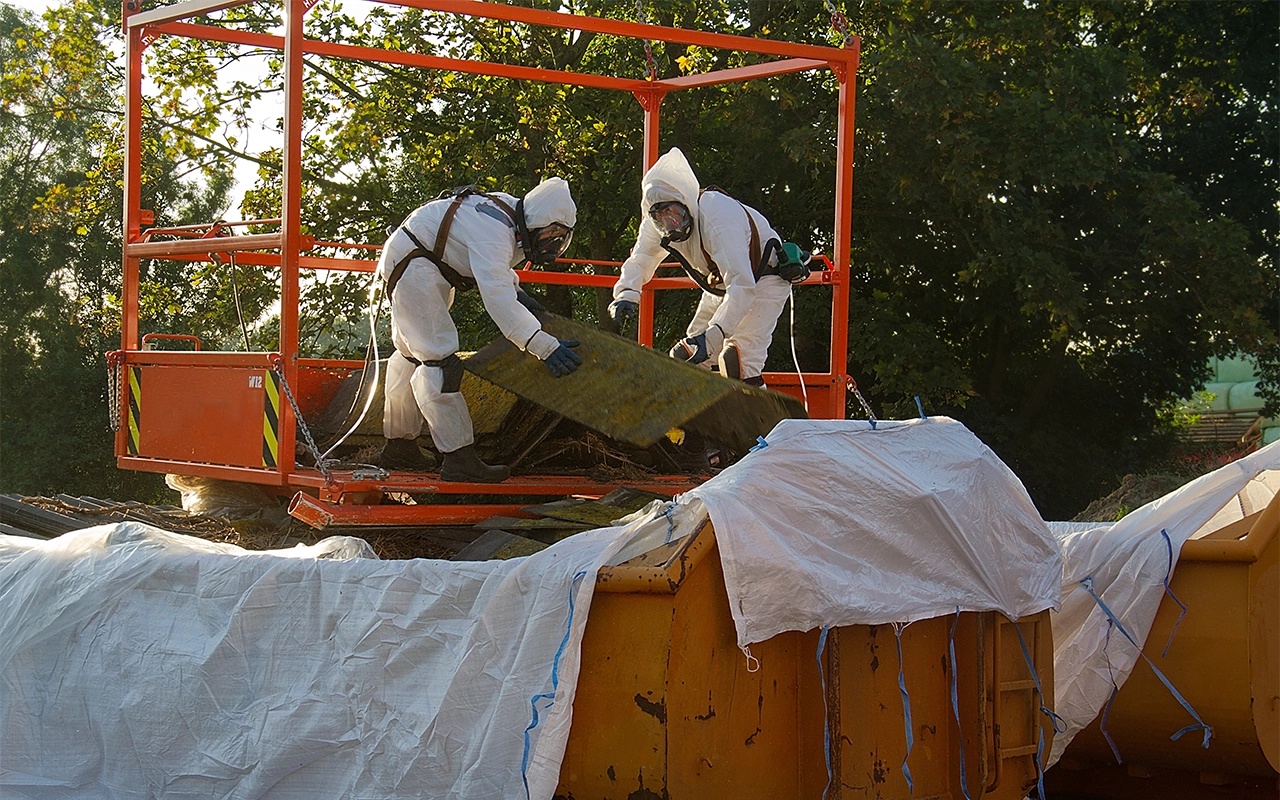 Deux hommes sur une plateforme métallique habillé en combinaison et masque en train d'intervenir sur un chantier contenant de l'amiante, ils trient l'amiante dans des sacs de tri spécifique à ce matériaux